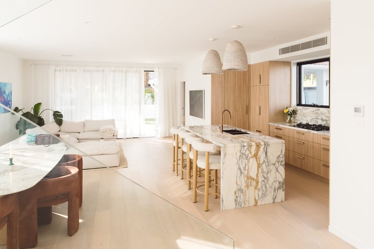 Modern open-plan kitchen and living room with wooden cabinetry, marble island, bar seating, and natural light from large windows
