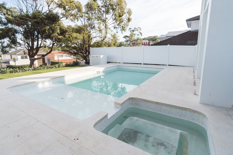 Modern backyard with rectangular swimming pool and spa, white concrete deck, and white privacy fence surrounded by trees
