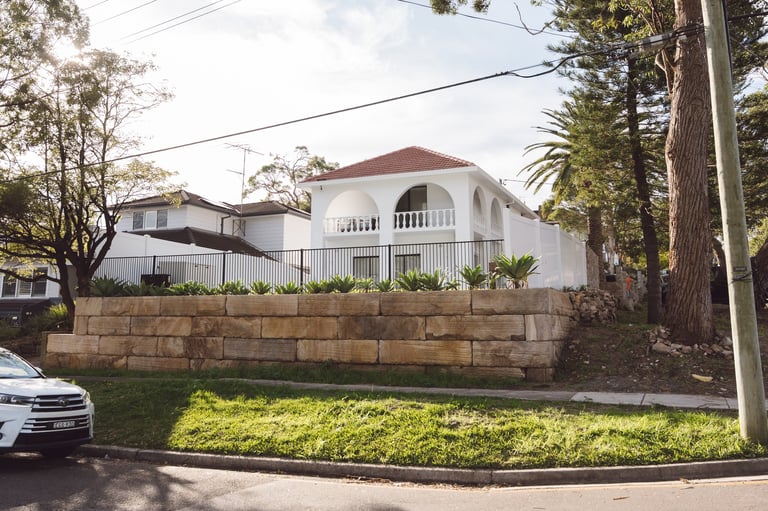 White Mediterranean-style house with red tile roof, stone retaining wall, and manicured lawn on a residential street