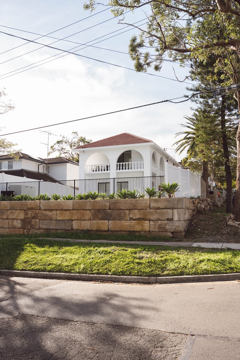 White elevated house with arched balcony and red roof behind a stone retaining wall with manicured lawn and tall trees