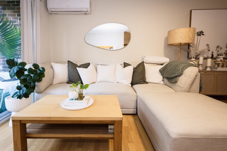 Modern living room with cream L-shaped sectional sofa, wooden coffee table, oval mirror, and decorative plants and lamp.
