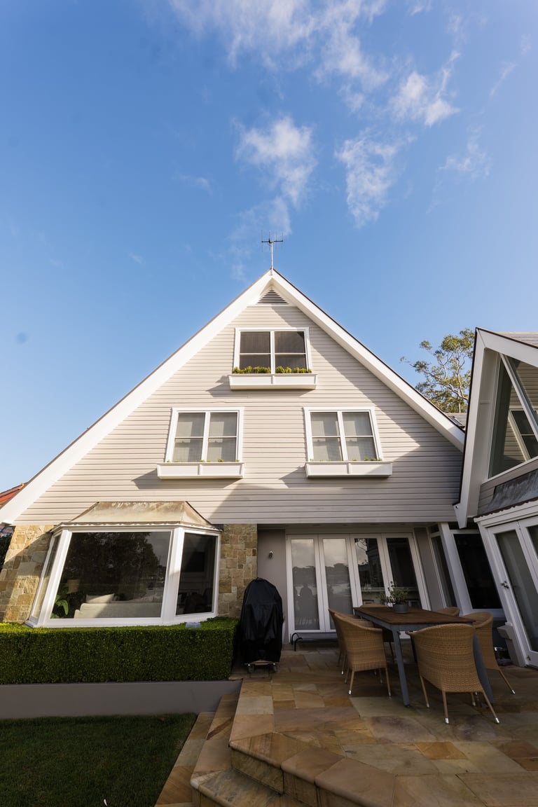 White A-frame house with pitched roof, cross on peak, multiple windows, and patio area with outdoor furniture under clear blue sky