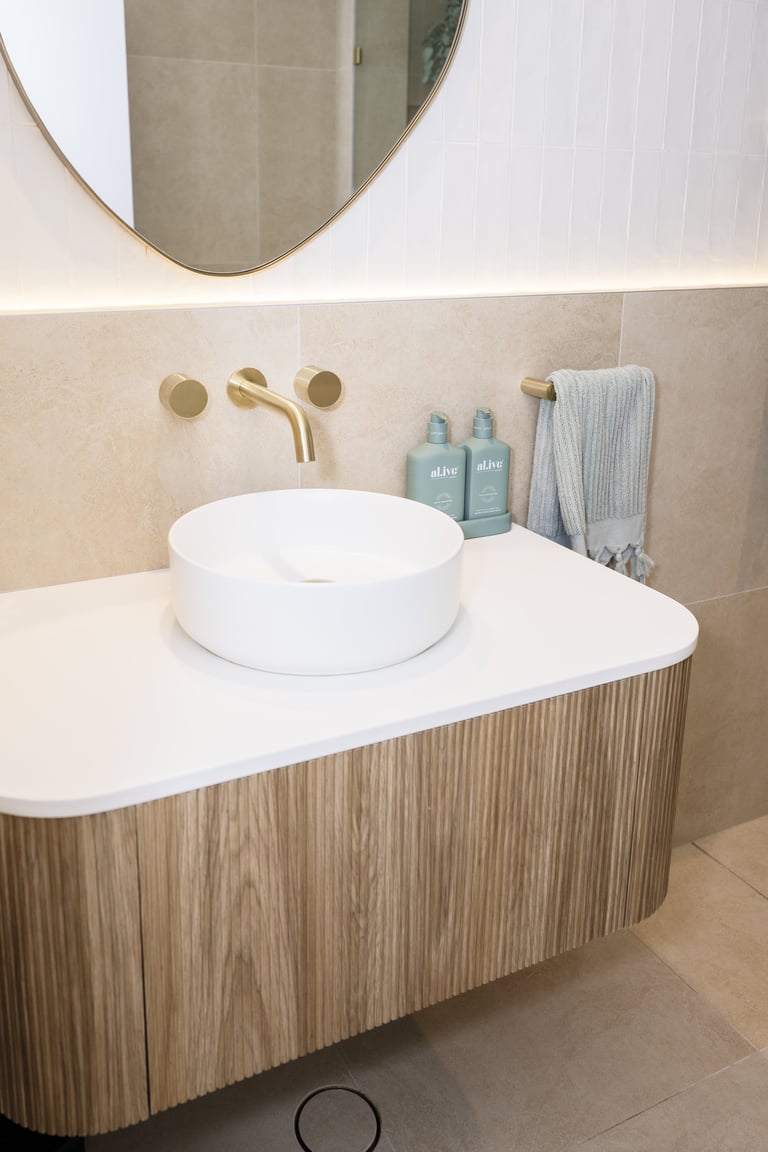 Modern bathroom vanity with white round vessel sink, wood-grain cabinet, brass wall-mounted faucet, round mirror, and shelf with toiletries