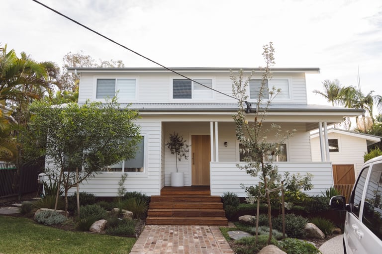 White two-story house with wooden entry deck, brick walkway, green landscaping, and palm trees
