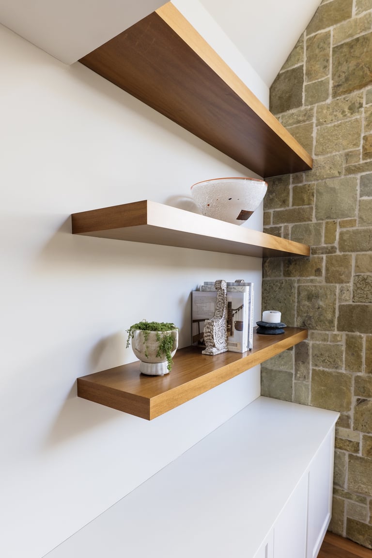 Three wooden floating shelves mounted on white wall next to stone brick, displaying decorative items and plants