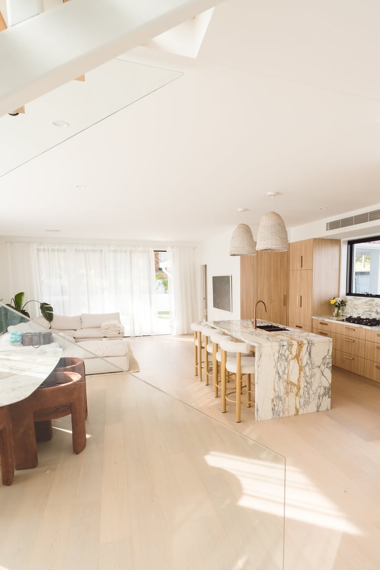 Modern open-plan kitchen and living room with marble island, wooden cabinetry, pendant lights, and cream tones throughout