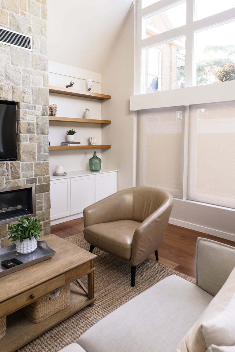 Modern living room with stone fireplace, wooden shelves, tan leather armchair, and large windows with natural light