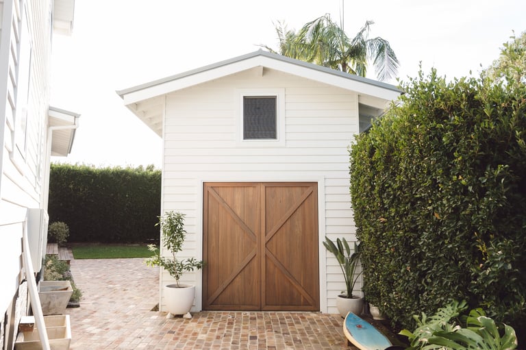 White garage with brown wooden doors surrounded by green hedges and potted plants on a brick patio