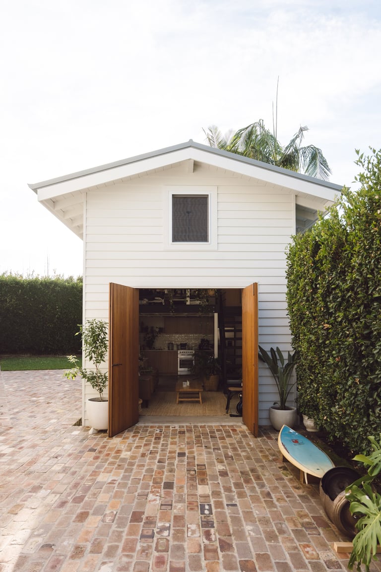 Modern white backyard studio with open wooden double doors, brick patio, and green hedges on sides