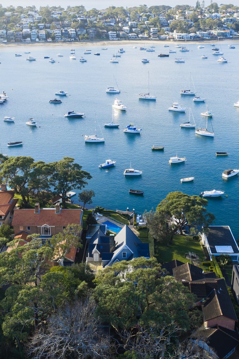 Aerial view of blue-roofed waterfront house surrounded by numerous moored sailboats in calm blue harbor water with tree-lined residential neighborhood behind