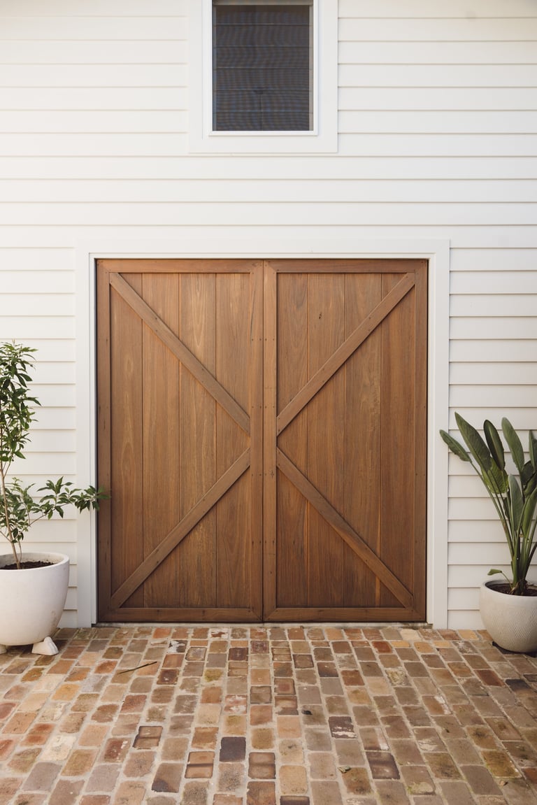 Wooden barn doors with X-pattern braces on white siding, flanked by potted plants on brick patio