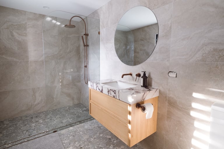 Modern bathroom with light wood vanity, oval mirror, marble tile walls, walk-in shower with glass enclosure, and brass fixtures