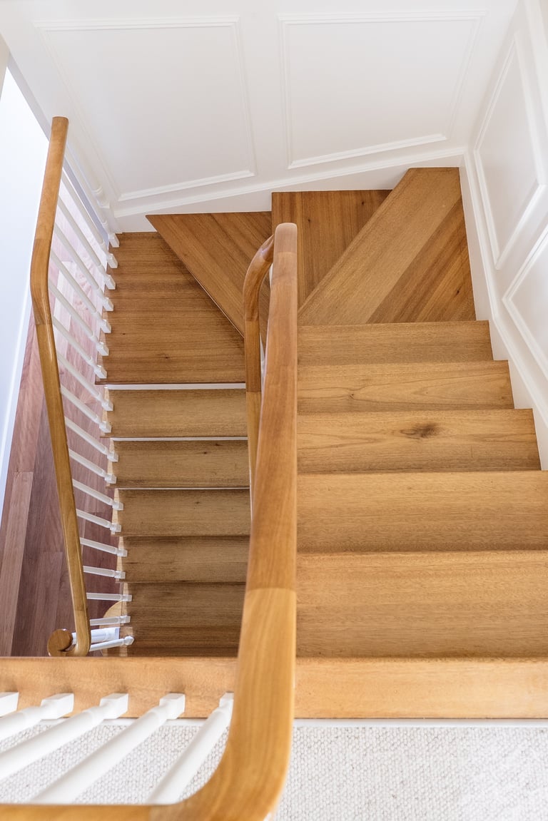 Wooden staircase with white walls and wooden handrail, viewed from above looking down at multiple landings and steps.