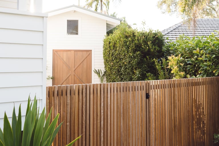 Wooden fence with a brown barn door and small window, with a white house and green hedges visible behind it