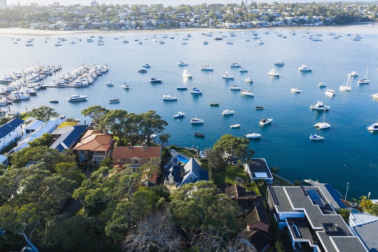 Aerial view of a coastal bay with numerous sailboats anchored in blue water, residential buildings and trees on a forested headland, residential neighborhood visible across the water