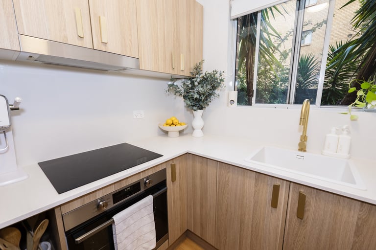Modern kitchen with light wood cabinets, white countertops, black stovetop, and tropical plants visible through windows