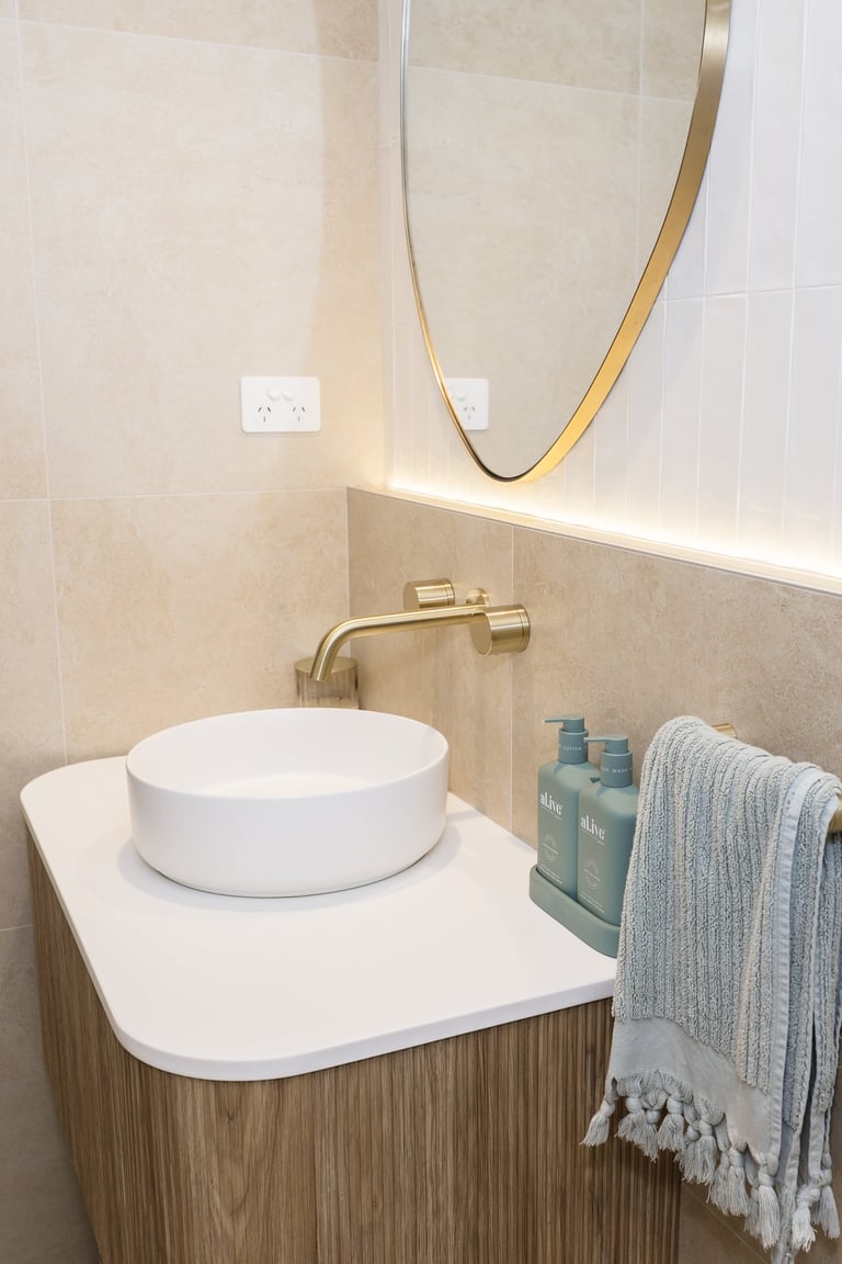 Modern bathroom vanity with white ceramic round sink, brass faucet, wooden cabinet, oval mirror, and towel rack