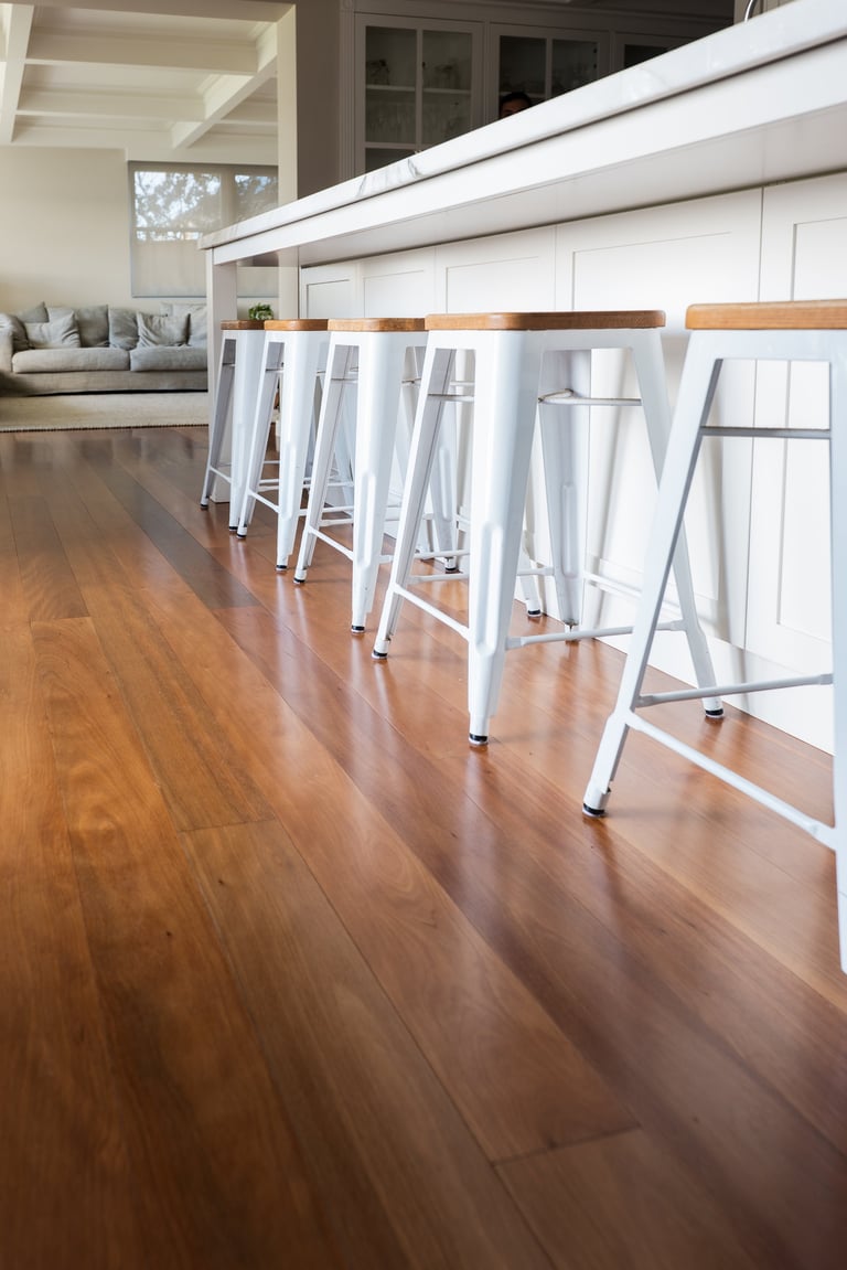 Modern kitchen with white metal bar stools with wood seats at long counter, hardwood flooring, and open seating area in background