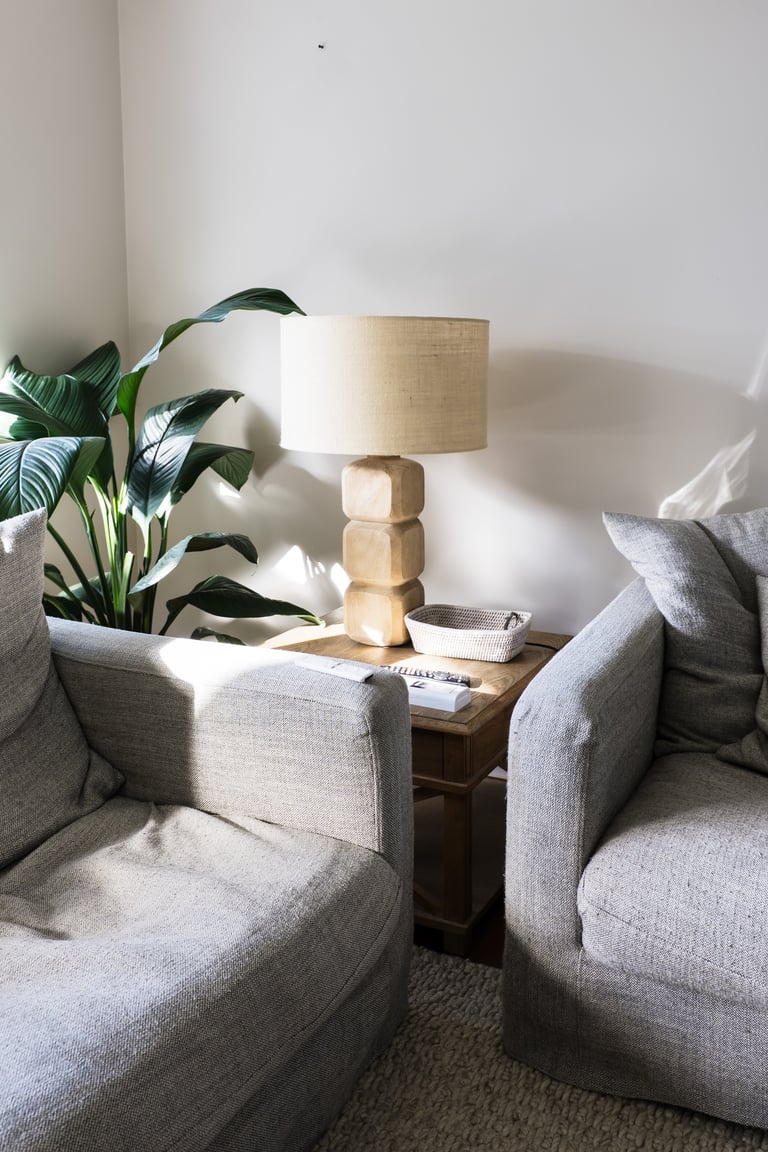 A cozy seating area with two gray upholstered armchairs facing a wooden side table with a stacked wooden base lamp and potted plant.
