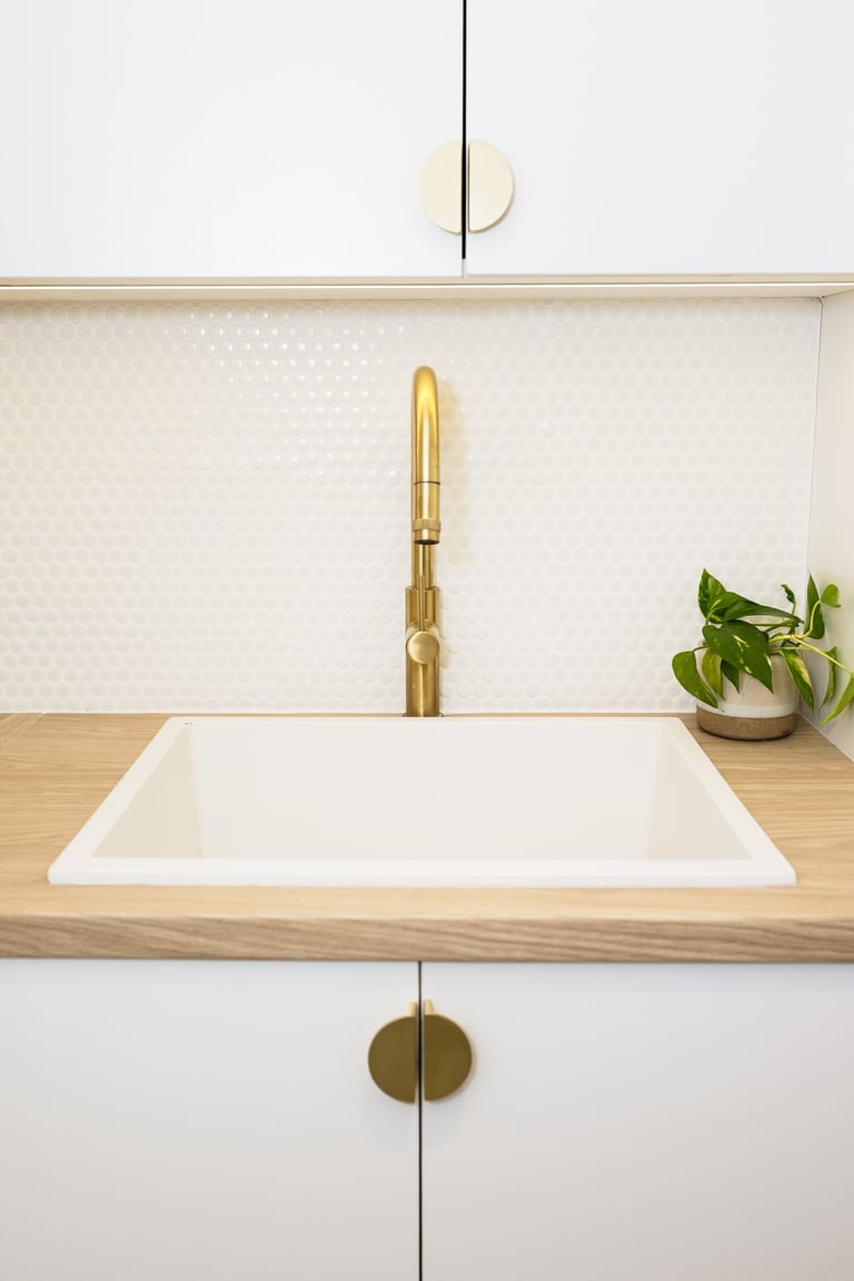 Modern minimalist kitchen sink with brass faucet, white tile backsplash, wooden countertop, potted plant, and gold hardware details