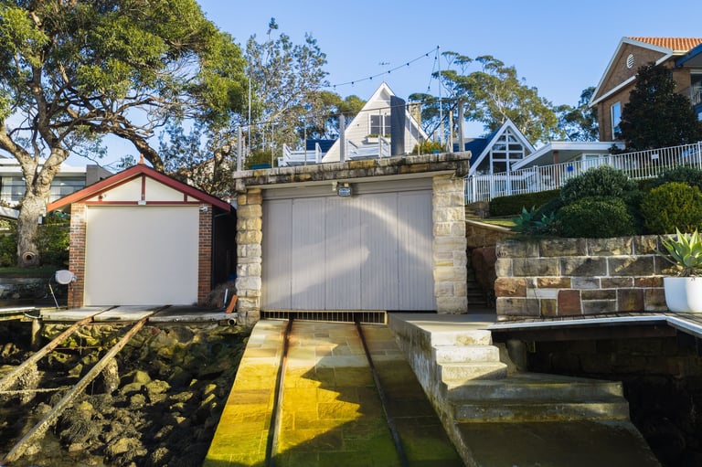 Two boat garages with white roller doors beside a waterway with moored boats, surrounded by residential houses and trees.