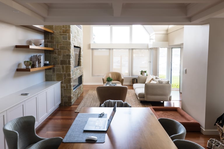 Modern open-concept living space with wooden desk in foreground, brick fireplace, white cabinetry, comfortable seating area, and large windows overlooking outdoor space