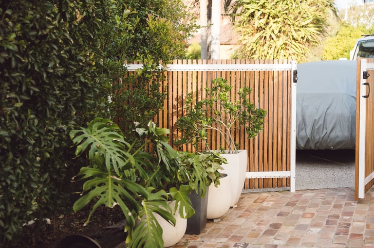 Modern patio with wooden fence gate, potted plants, and curved white planter, adjacent to a covered outdoor structure