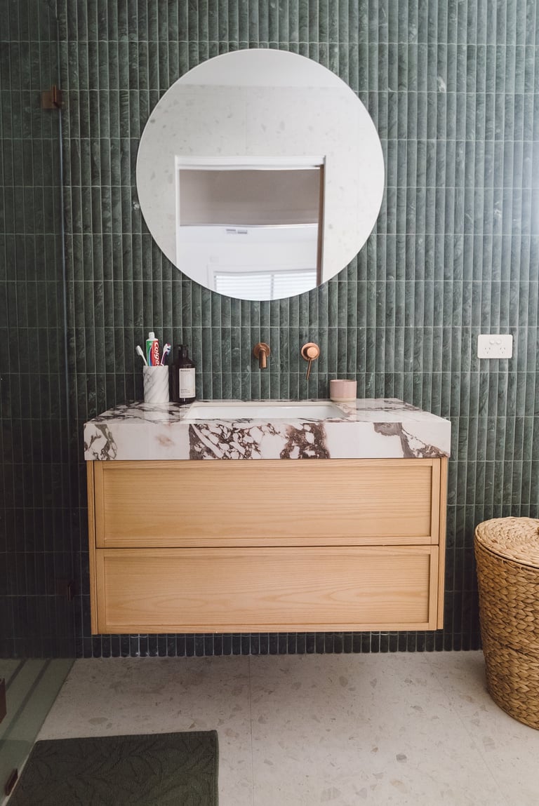 Modern bathroom vanity with wooden drawers, marble countertop, round white mirror, green tile wall, and woven basket accessory