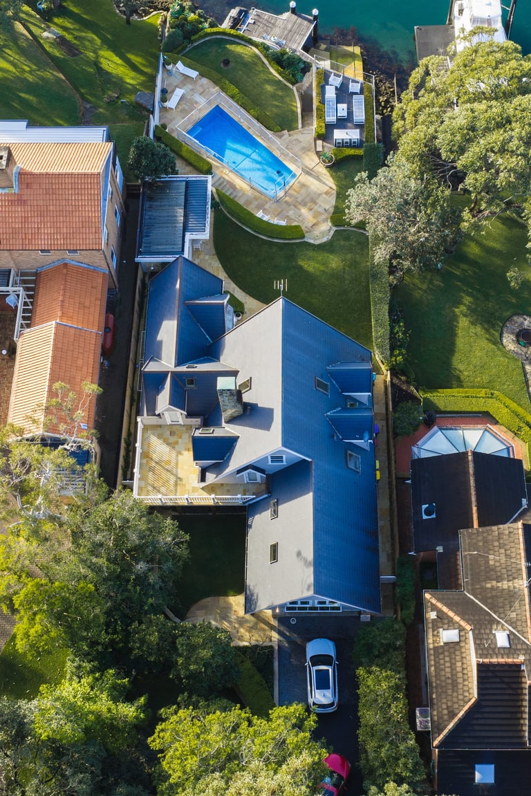 Aerial view of a waterfront property with blue roof, swimming pool, surrounding green grounds and moored boats