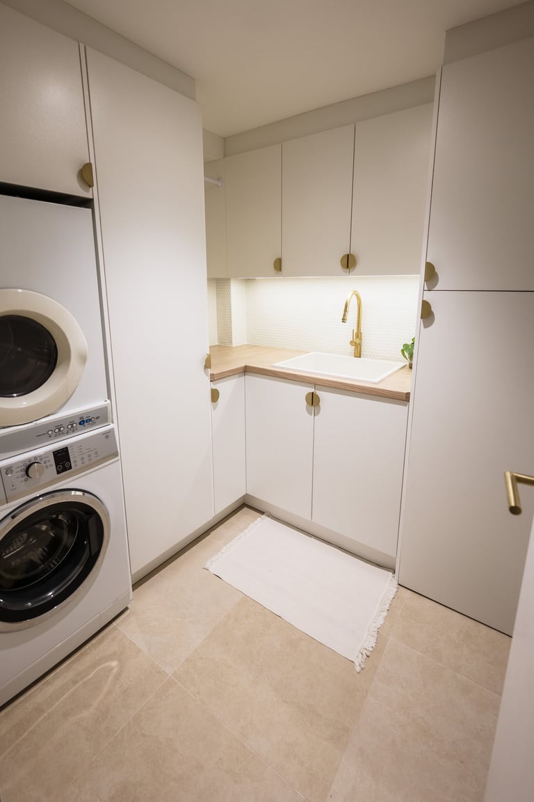 Modern laundry room with stacked washer and dryer on left, white cabinetry, sink with gold faucet, and light wood countertop