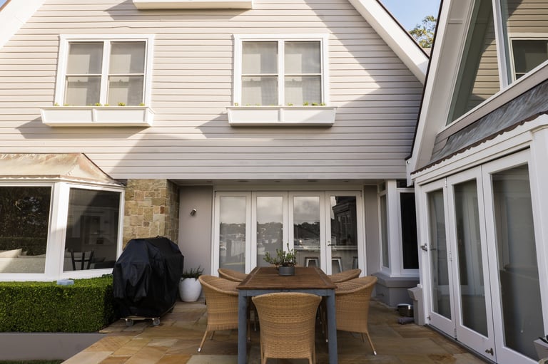 Modern white house with patio area featuring wooden dining table, chairs, and BBQ grill under covered space