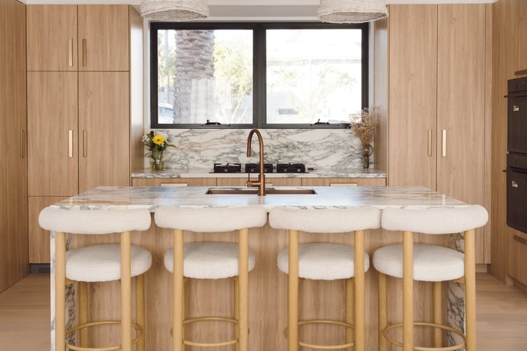 Modern kitchen with wooden cabinetry, marble countertop island with four bar stools, and large window above stainless steel sink