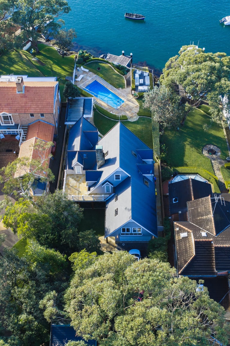 Aerial view of a blue and white waterfront house with manicured lawn, swimming pool, and moored boats on turquoise water