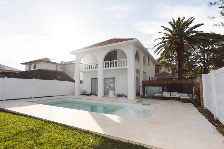 Modern white villa with swimming pool, manicured lawn, and palm tree surrounded by white fencing