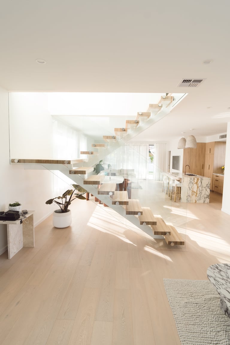 Modern minimalist interior with floating wooden staircase, light hardwood floors, white walls, and natural sunlight streaming through windows