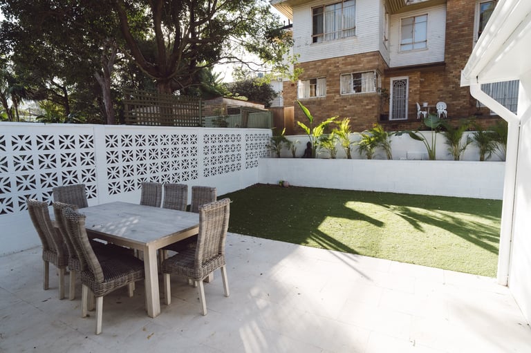 Modern backyard patio with dining table, chairs, and artificial grass surrounded by white fence and brick house