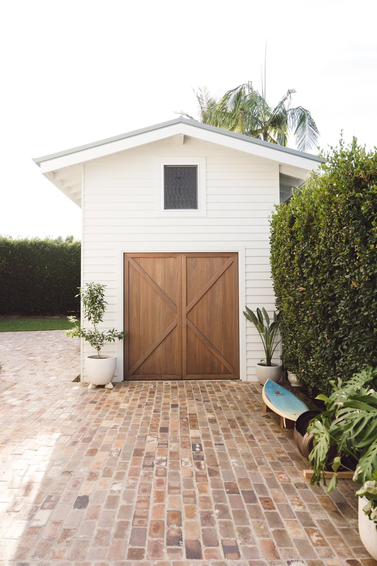 White modern garage with large wooden double doors and brick driveway, surrounded by greenery and potted plants