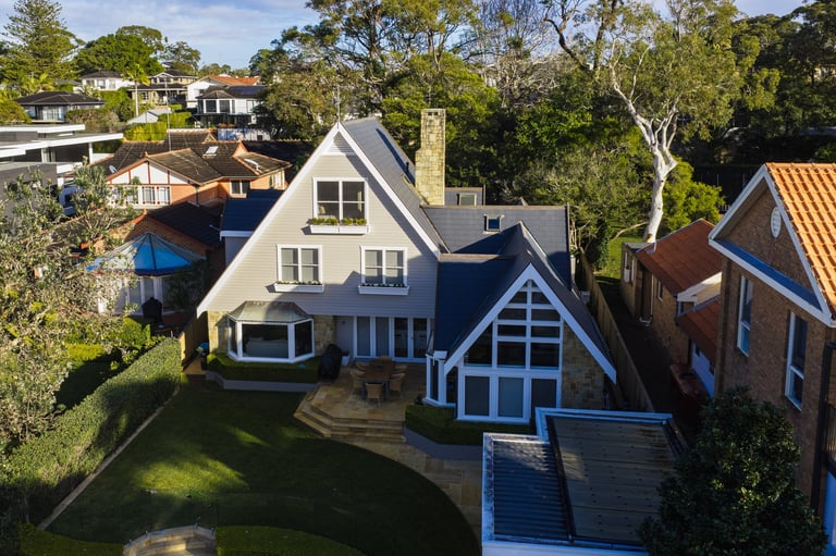 Aerial view of a blue and white residential house with distinctive Tudor-style architecture and manicured lawn surrounded by trees