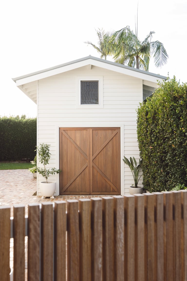 Modern white garden shed with brown wooden barn doors, potted plants, and green hedges beside a wooden fence