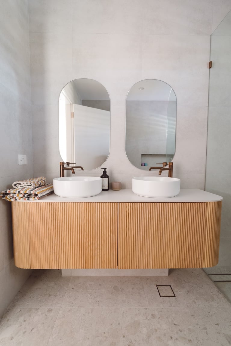 Modern bathroom vanity with wooden ribbed base, white countertop, two round white sinks, and two oval mirrors with bronze fixtures
