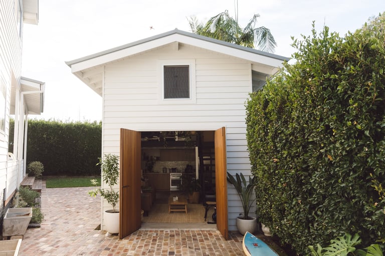 Modern white garden studio with wooden doors, brick patio, and green hedges on either side in residential backyard setting