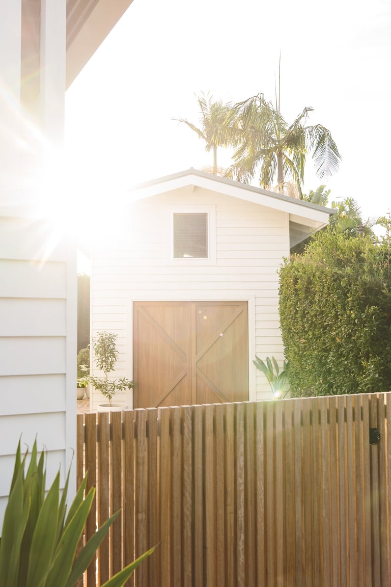 Modern white two-story house with wooden garage doors and fence, tropical plants, bright sunlight