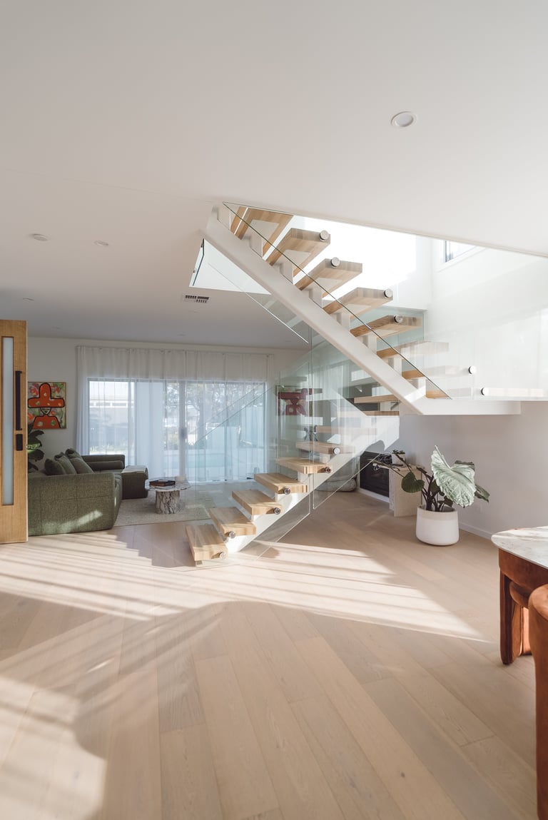 Modern open-plan living space with floating wooden staircase, light wood flooring, and floor-to-ceiling windows overlooking trees